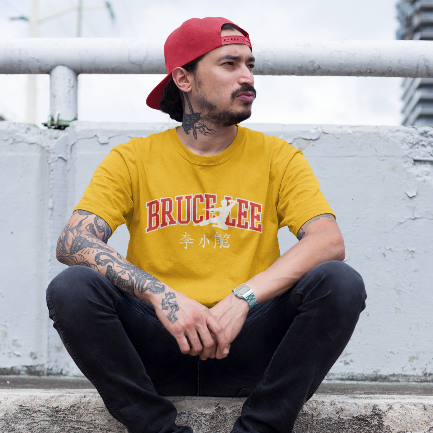 Man in yellow Bruce Lee Varsity Flying Man t-shirt and red cap by white railing